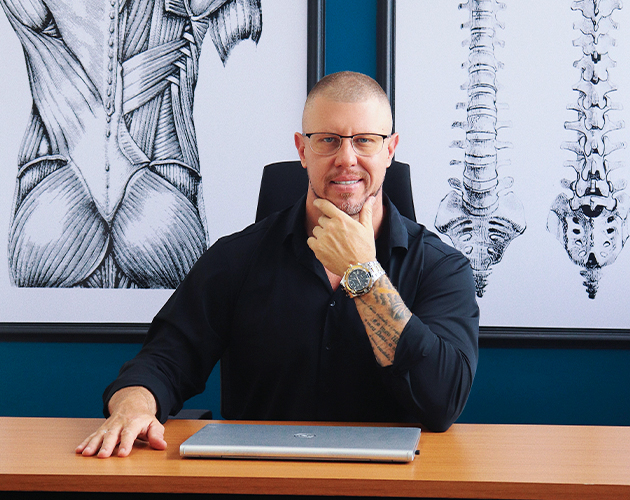 Dr Ronaldo Nigro, fisioterapeuta especializado em coluna vertebral e dores crônicas em consultório, ao lado de ilustração anatômica da coluna.